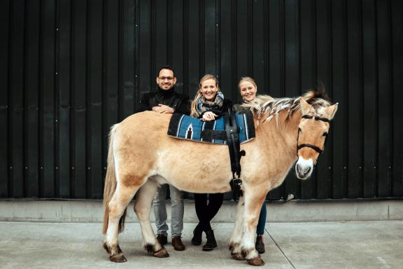 Reittherapie Ivenack Drei lächelnde Therapeuten stehen hinter einem Pferd und schauen über den Rücken.