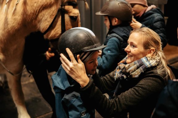 Reittherapie Ivenack Eine Frau zieht einem Kind einen Reithelm an, beide sind fröhlich und gespannt.