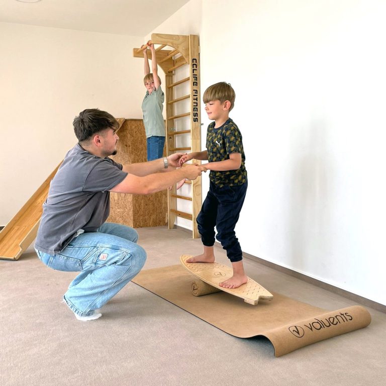 Ergotherapeutische Praxis Robert Steiger Ein Mann stützt einen Jungen bei einer Balanceübung auf einem Board, im Hintergrund turnt ein Junge an einer Sprossenwand.