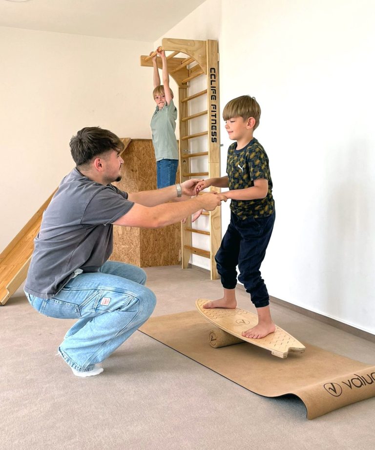 Ein Mann stützt ein Kind auf einem Balanceboard, im Hintergrund turnt ein Junge an einer Sprossenwand.