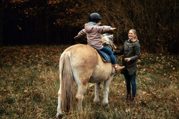 Reittherapie Ivenack Ein Kind mit einem Reithelm auf, sitz auf einem Pferd und spreizt die Arme seitlich, eine Therapeutin steht davor und ermutigt es.